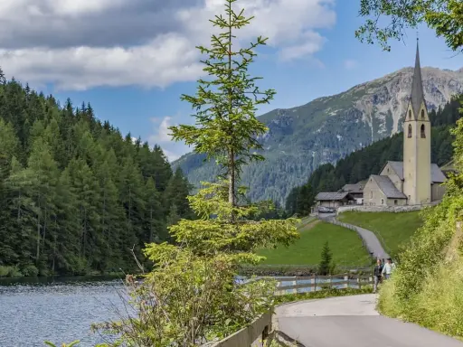 Passeggiata sul lago di Valdurna, con boschi rigogliosi, una chiesa alpina e montagne sullo sfondo.