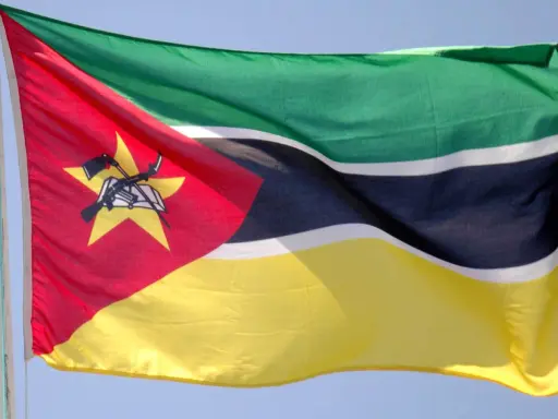 Flaga Mozambiku: zielono-czarno-białe pasy, czerwony trójkąt z żółtą gwiazdą, karabin AK-47 i książka.