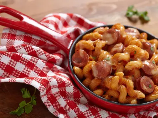 Pyszne spaghetti z kiełbasą w sosie pomidorowym, podane w czerwonej patelni, obok czerwono-białej kratkowanej serwetki.