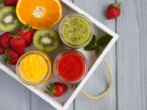 Bandeja con fresas, rodajas de kiwi, naranja y tres tarros de **puré de frutas** de colores vibrantes: verde, amarillo y rojo.