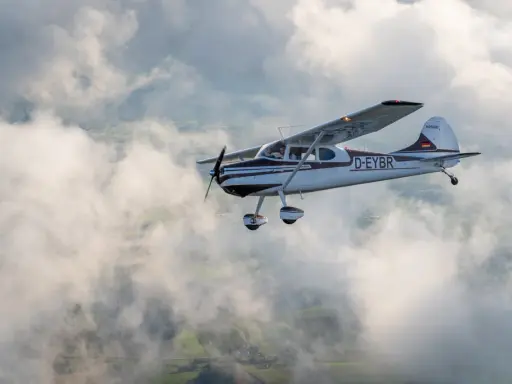 Eine braun-weiße Cessna 170 fliegt durch die Wolken über grünen Feldern.