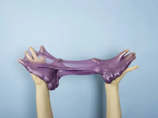 Manos estirando un brillante slime casero morado con purpurina sobre fondo azul.