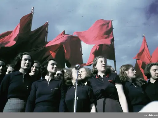 En grupp kvinnor deltar i ett 1 maj tåg, med röda fanor vajande i bakgrunden.