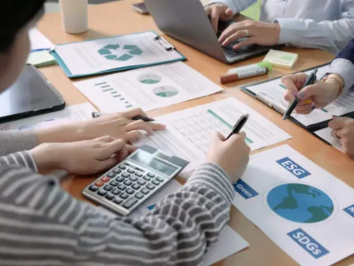 Des mains travaillent sur des documents financiers, une calculatrice et un ordinateur portable. L'ergonomie organisationnelle est au cœur de cette réunion axée sur le développement durable.