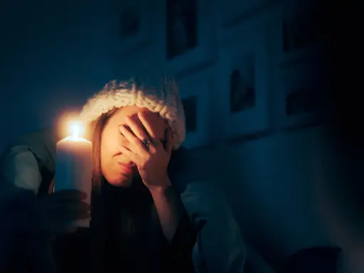 Kobieta w czapce trzyma świecę, zasłaniając twarz. To skutki przerwania dostaw energii elektrycznej do urządzeń o kluczowym znaczeniu.