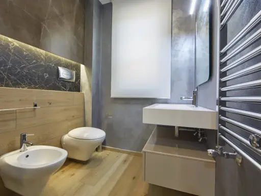 Bagno moderno con bidet, WC sospeso, lavabo e scaldasalviette cromato. L'illuminazione a LED sotto il marmo crea un'atmosfera elegante, rispettando l'altezza minima soffitto.