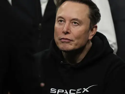 Elon Musk w bluzie SpaceX, zamyślony, w otoczeniu innych osób.
