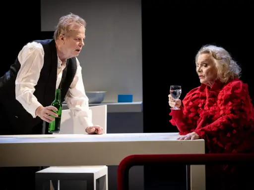 Krister Henriksson i en dramatisk scen. Han håller en grön flaska, medan en kvinna i rött med ett glas vatten ser på. Scenen påminner om hans roller i filmer och tv-program.