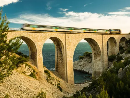 Ein Zug rast über eine Bogenbrücke, die Teil des Bahnnetz Frankreichs ist. Das Meer glitzert unter den Bögen.