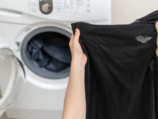 Manos sostienen ropa negra con manchas blancas, cerca de una lavadora abierta. ¿Por qué la ropa negra sale con manchas blancas?