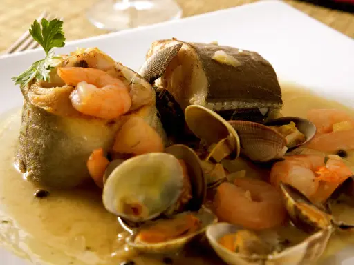 Delicioso abadejo a la donostiarra con gambas y almejas en una salsa dorada, adornado con perejil fresco.