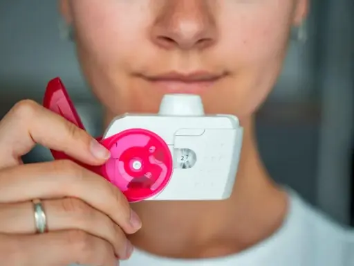 Nahaufnahme einer Person, die einen Pulverinhalator hält, bereit zur Anwendung. Der Inhalator ist weiß mit pinken Akzenten und zeigt die Dosis "27".