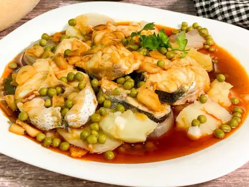 Merluza a la gallega con patatas, guisantes y ajo frito en una salsa rojiza.