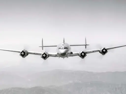Ein vier-motoriger Propellerflieger mit markantem Leitwerk fliegt über bergiger Landschaft.