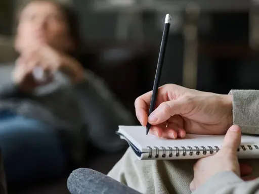 Psycholog pomaga zrozumieć siebie i swoje problemy. Na zdjęciu widać rękę terapeuty piszącą w notatniku, podczas gdy pacjent leży na kanapie.
