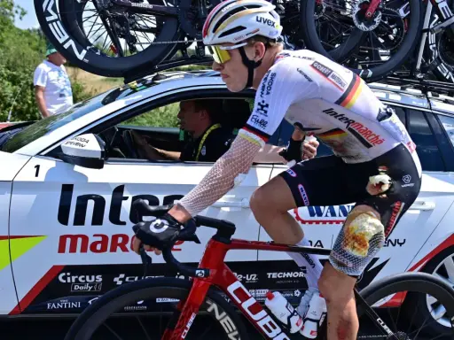 Radfahrer Georg Zimmermann mit aufgeschürftem Knie fährt neben dem Begleitfahrzeug des Teams Intermarché-Wanty.