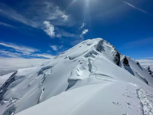 Śnieżna trasa na Mont Blanc, z widocznymi szczelinami lodowcowymi i śladami wspinaczy. Błękitne niebo z chmurami.