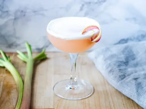 En rosa rabarber sour-drink med skum och en rabarberdekoration. Två stjälkar rabarber ligger bredvid.