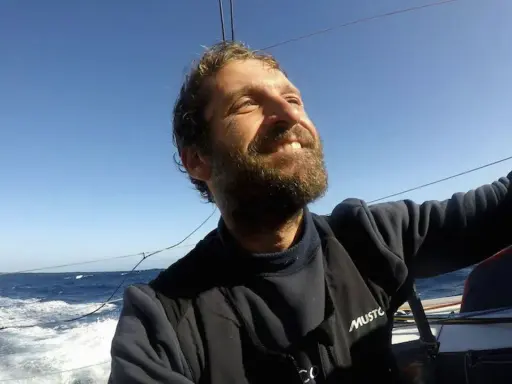 Un homme de mer souriant, le visage tourné vers le ciel bleu, navigue sur un voilier. L'océan agité et le soleil radieux témoignent de son aventure.