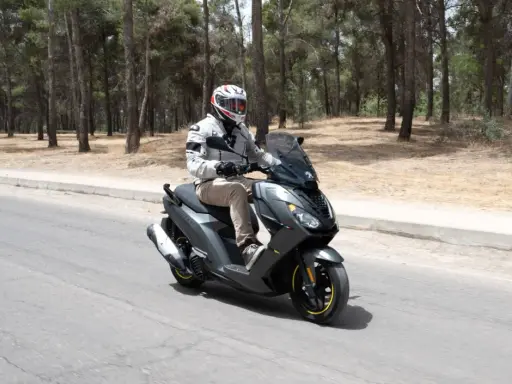 Hombre en casco y chaqueta conduce un triciclo de motor gris por una carretera rodeada de pinos.