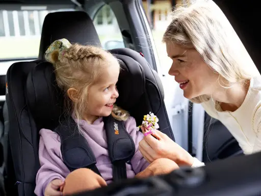 Uśmiechnięta mama daje córeczce w foteliku samochodowym bukiecik kwiatów. To miły test plus dla małej pasażerki.