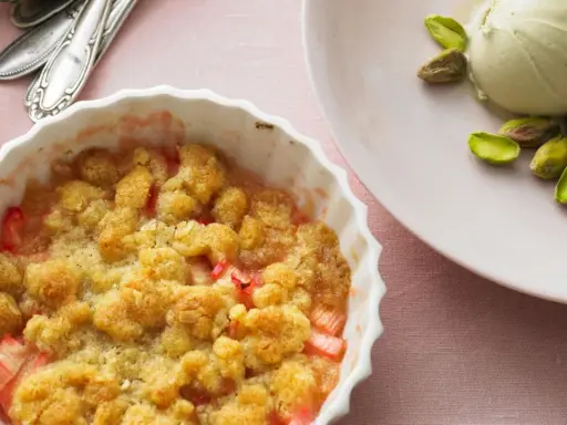 Rhabarber-Crumble in weißer Auflaufform, daneben eine Kugel Eis mit Pistazien. Ein köstliches Rhabarber Dessert.