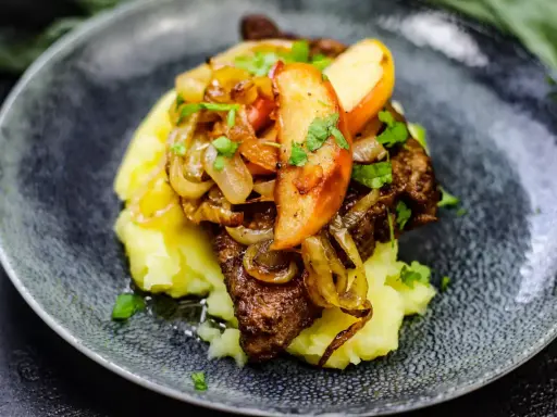 Gedünstete Leber mit Apfelspalten, Zwiebeln und Kartoffelpüree auf einem dunklen Teller.