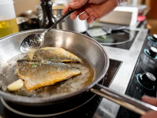 Un chef cocina pescado en una sartén de acero inoxidable, bañándolo con aceite caliente.