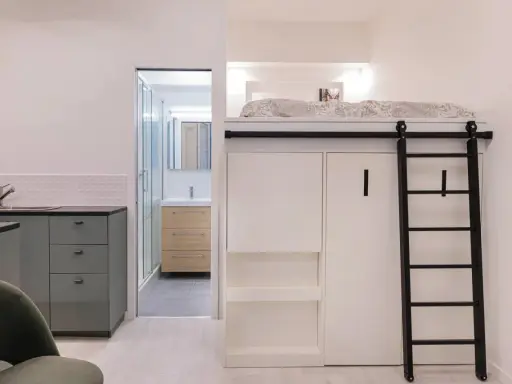 Un appartement fonctionnel avec lit mezzanine, échelle noire, cuisine grise et salle de bain visible.