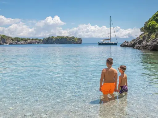 Due bambini esplorano le acque cristalline del Cilento, tra le sue spiagge perfette per famiglie. Una barca a vela all'orizzonte completa il quadro idilliaco.
