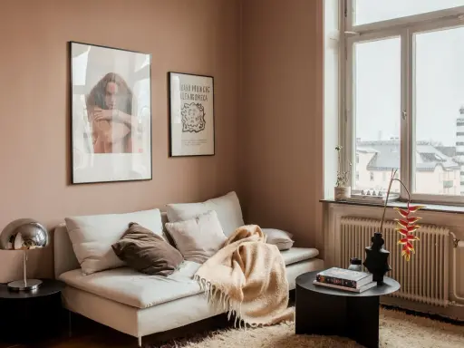 Un divano bianco accogliente con cuscini e una coperta color **rosa pastello** su un tappeto soffice. Alle pareti, due quadri e una finestra con vista.