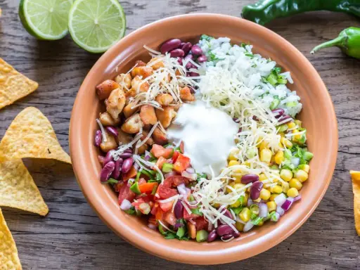 Bunter Taco Salat mit Hähnchen, Reis, Bohnen, Mais, Tomaten, Käse und Sauerrahm. Tortilla-Chips und Limetten daneben.