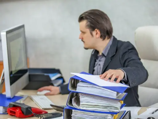 Mężczyzna w garniturze pracuje przy biurku, przeglądając dokumenty. Przed nim stos segregatorów, komputer i czerwony telefon. To obraz codziennego postępowania administracyjnego.
