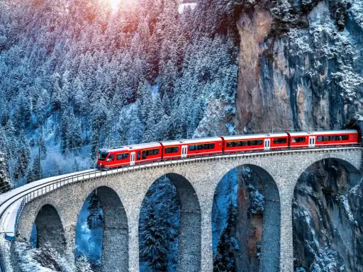Un treno rosso attraversa un viadotto innevato, parte del suggestivo treno Bernina percorso, tra alberi coperti di neve e rocce imponenti.
