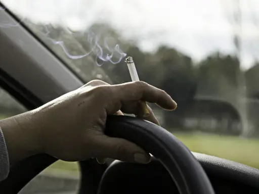 Mano sujetando un cigarrillo encendido sobre el volante. El humo se eleva mientras se puede fumar conduciendo.