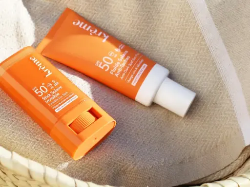 Deux produits solaires, un stick et un fluide, sont posés sur une serviette rayée. Le bouton du stick est visible, prêt à protéger du soleil.