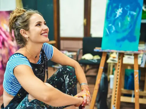 Une artiste rêveuse, les mains tachées de peinture, s'interroge sur comment devenir artisan d'art. Son atelier est rempli de toiles colorées.