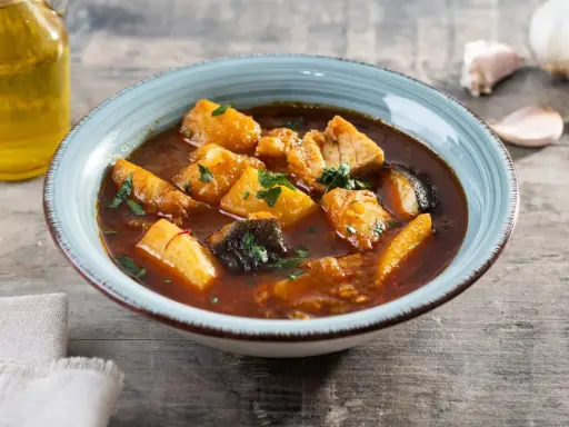 Un tazón de patatas con bacalao en salsa, decorado con perejil. Al lado, una botella de aceite de oliva y ajos.