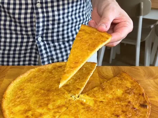 Una fetta di farinata di ceci dorata e croccante viene sollevata da una teglia rotonda su un tagliere di legno.