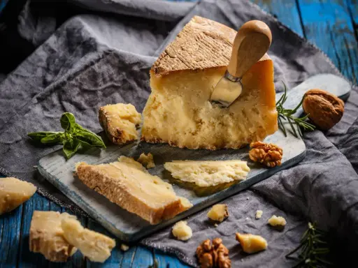 Kawałek parmezanu z nożem, okruchy sera, orzechy i bazylia. Czy parmezan można mrozić? Tak, można go mrozić, choć może stracić nieco na konsystencji.