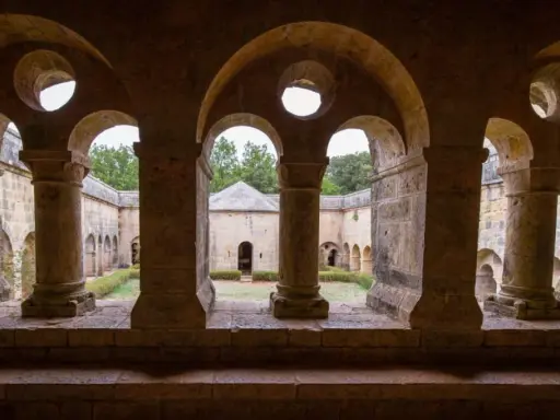 Vue à travers les arches de l'abbaye du Thoronet, un cloître en pierre avec un jardin verdoyant et des bâtiments anciens.