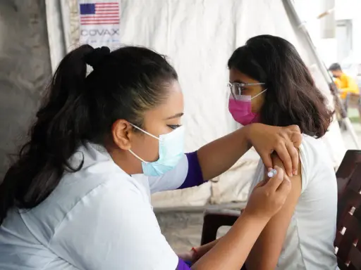 En sjuksköterska ger en vaccination i Nepal. Bakom syns en banderoll med texten COVAX och en amerikansk flagga.
