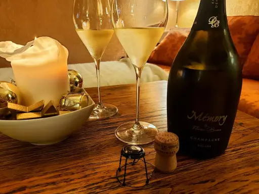 Zwei Gläser Champagner, eine Kerze und eine Flasche "Memory" auf einem Holztisch. Der Champagnergehalt ist hierbei nicht ersichtlich, aber die Stimmung ist festlich.