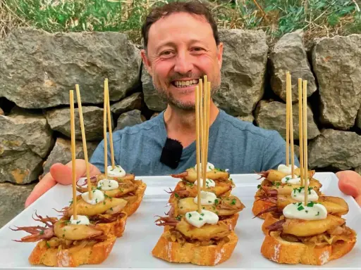 Hombre sonriente presenta cazuelitas de aperitivo con calamares y alioli sobre pan tostado, decoradas con brochetas.