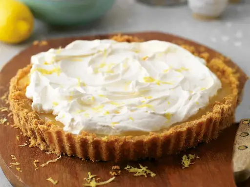 A lemon tart topped with whipped cream and lemon zest on a wooden board.