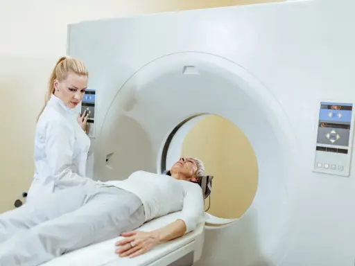 Kobieta w białej bluzie leży na łóżku rezonansu magnetycznego. Technik w białym fartuchu obsługuje panel sterowania.
