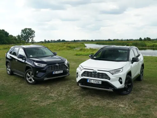 Dwa modele Toyota RAV4 2020, jeden czarny, drugi biały, stoją na trawie nad jeziorem.