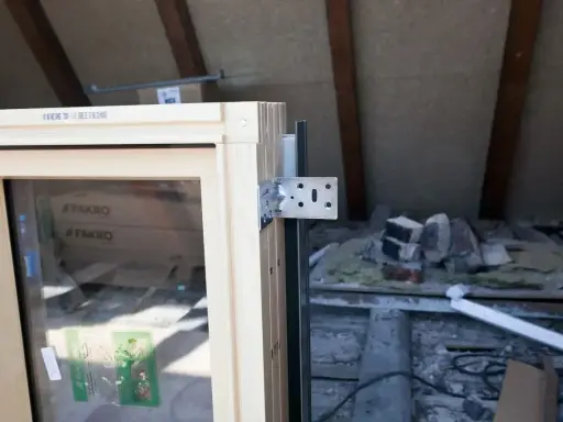 Drewniane okno z metalowym kątownikiem, gotowe do obróbki po montażu. W tle widać materiały budowlane i drewniane belki poddasza.