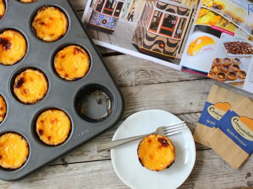 Pyszne, lekko przypieczone pastel de nata w blaszce i na talerzyku. Obok magazyn z widokiem na Lizbonę i tramwaje.