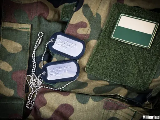 Dwa nieśmiertelniki na mundurze moro. Jeden z nich zawiera napis "MILITARIA.PL SHOOTING & OUTDOOR".